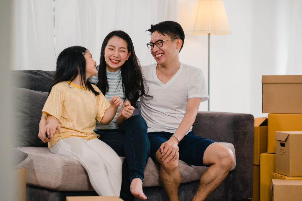 happy-asian-young-family-homeowners-bought-new-house-japanese-mom-dad-daughter-embracing-looking-forward-future-new-home-after-moving-relocation-sitting-sofa-with-boxes-together-scaled